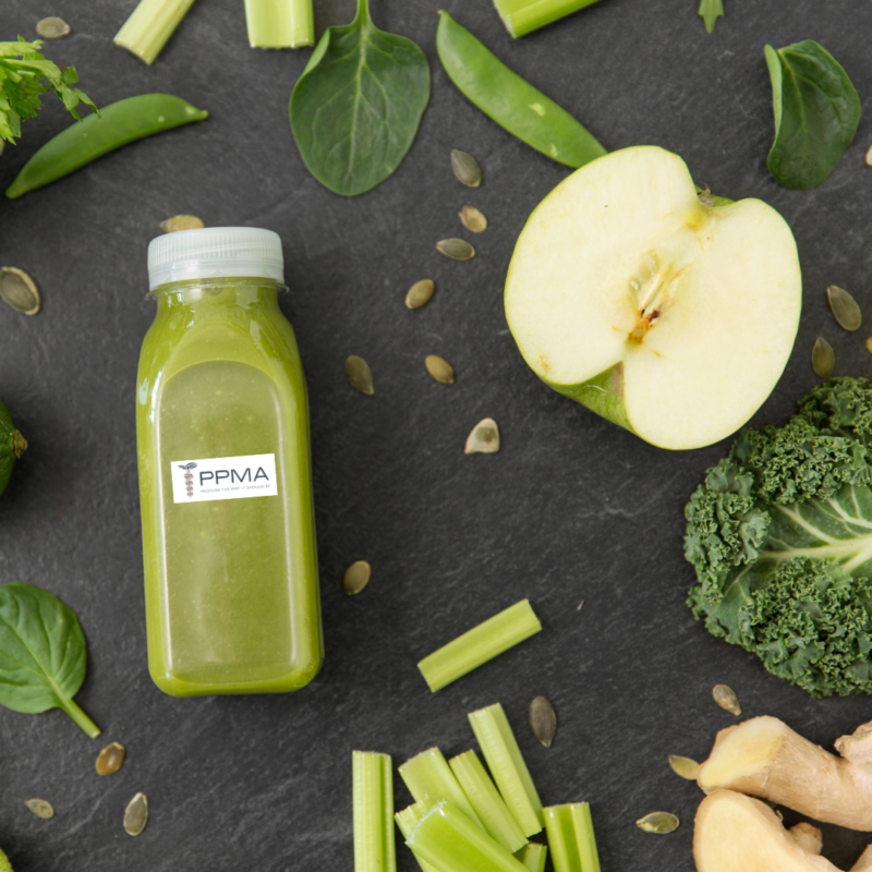 Image of clear jug of apple-kale smoothie surrounded by half green apple, celery, ginger, seeds and kale on a dark wood background+