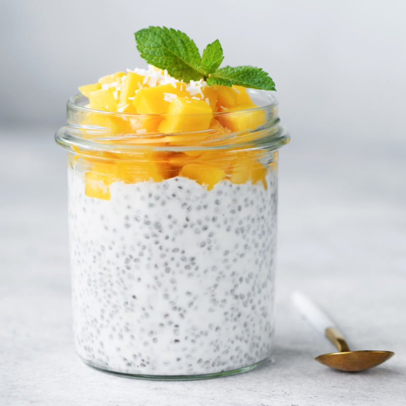 Image of Mango Chia Pudding in glass with fresh cut mango and sprig of mint on top, with a spoon to the side on white-gray back