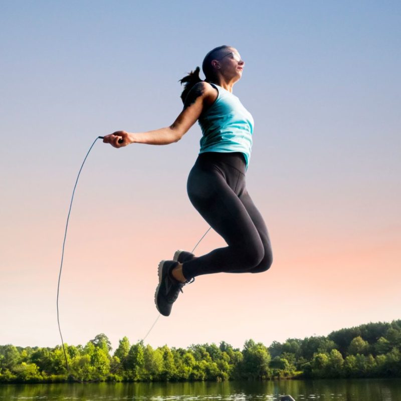 Image of a woman jumping rope with jump rope outdoors near body of water. She appears to be very high in the air. For PPMA exercise regularly
