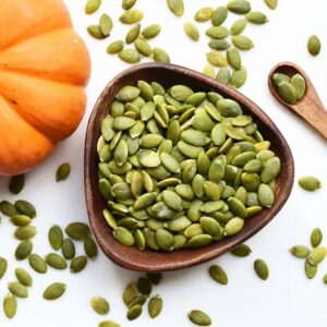 Image of raw pumpkin seeds in bowl, in wooden spoon, and scattered on white countertop. Part of whole, uncut pumpkin on side. Good source of zinc. Romana Brennan MS RD PPMA Private Physicians Medical Associates Concierge Medicine, Newport Beach, California