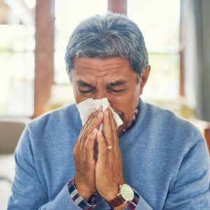 Image of mature adult wearing a blue sweater blowing their nose into a tissue. Prep for Flu Season NOW PPMA. Private Physicians Medical Associates