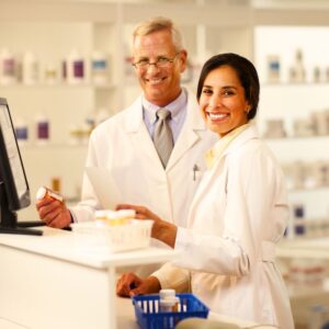 Image of two pharmacists, a middle aged, gray-haired man and a middle aged woman, standing in a pharmacy, smiling. PPMA. Private Physicians Medical Associates.