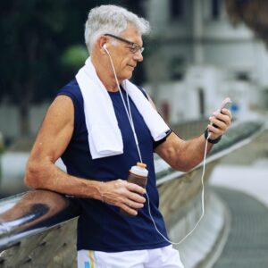 An image of a middle-aged man dressed in workout clothes, glasses, and earbuds, leaning against a rail as he checks his mobile phone. He's holding a bottle of water in his right hand. The Art of Aging Well.