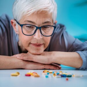 In this image, a woman with blue glasses and short, straight, silver hair is resting her chin on her hands, which are one atop the other on a table, as she looks at a scattering of prescription medications on the table.