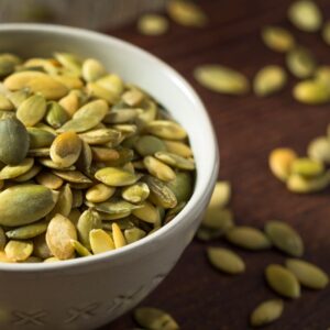 This image is a close-up of a white bowl holding raw pumpkin seeds, with more pumpkin seeds scattered around the bowl.