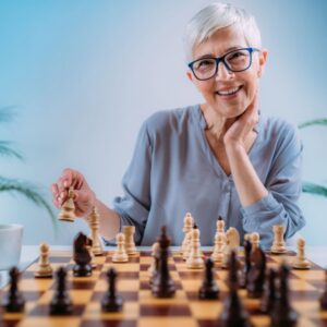 This is an image of a happy, smiling middle-aged woman with short silver hair, blue glasses and a blue shirt. She is playing chess, showing her cognitive functional abilities.