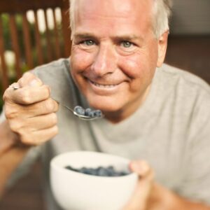 This is an image of a middle-aged man smiling as he eats blueberries with a spoon from a bowl.