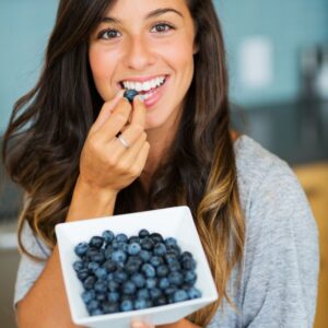 A young woman smiling as she eats blueberries from a white bowl.