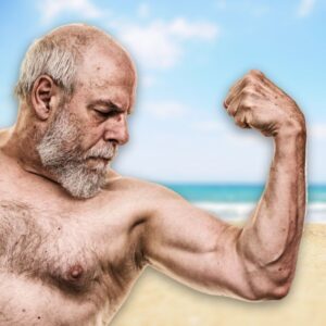 An image of a middle-aged or senior man flexing his left arm and showing muscles with the beach and ocean behind.