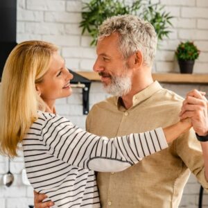 An attractive middle-aged couple is dancing in their kitchen and smiling at each other.