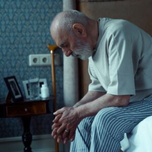 An image of a senior man sitting on the edge of his bed looking sad. Mental health.
