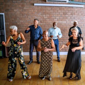 Middle aged and senior friends dancing together on a wooden floor with a red brick background. 