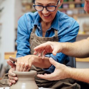 A smiling older woman wearing blue glasses and a blue denim shirt is being taught to use a clay wheel.