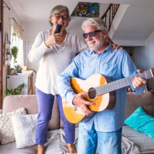 A middle-aged or senior couple at home smiling and making music - the man playing guitar, the woman singing.