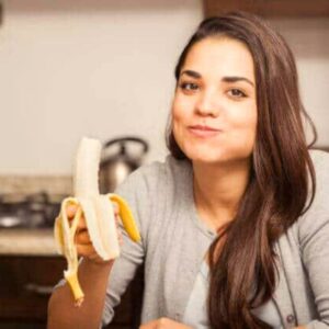 An image of a young woman chewing a bite of banana as she holds the banana in her hand. Gut health.