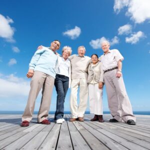 An image of five senior or middle-aged friends, 3 men and 2 women, smiling at the camera on a dock with the sea and sky behind them.