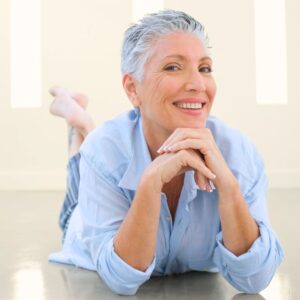 An image of a lovely middle-aged woman with short silver and gray hair, laying on her belly with her chin resting on her hands, smiling at the camera. The Power of Preventive Care