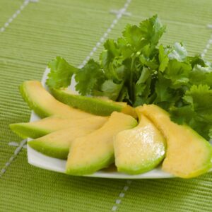 A plate of sliced avocado on a white plate and a cilantro garnish, atop a green woven tablecloth.