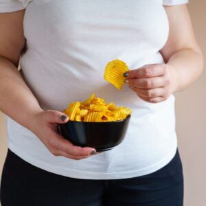 An image of an overweight person wearing a white tee shirt and black pants holding a black bowl of potato chips, one chip in her hand.