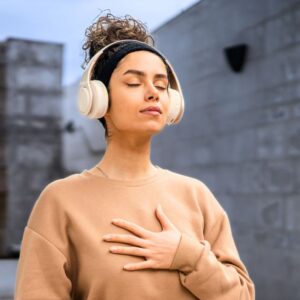An image of a woman wearing white headphones with her left hand on her chest, eyes closed, as she meditates.