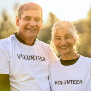 A senior or middle-aged couple posed in matching white tee shirts that say "VOLUNTEER" on them in black letters. Both are smiling with nature behind them. Retiring: Is It Time?