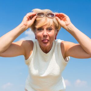 An image of a middle-aged woman or senior woman leaning towards the camera as she smiles, playfully sticking her tongue out, with a blue sky behind her.