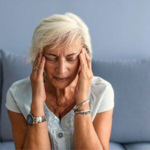 A middle-aged or senior woman seated and holding her temples with her fingers. Her eyes are closed and she has a headache. Hydrate For Your Health