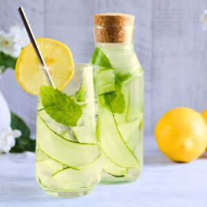 An image of a glass bottle and glass jar, both containing water with thinly sliced cucumber, mint leaves, and lemon slices, with a whole lemon to the side.