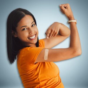 A woman dressed in an orange short-sleeved top making a muscle pose pointing with her left hand to a bandage on her right arm. Influenza or flu vaccine