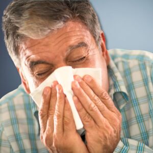 An image of a man in a blue checked shirt blowing his nose in a tissue.