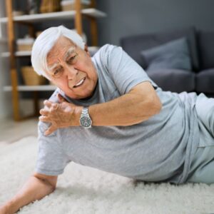 An image of a senior man grimacing in pain after falling to the floor in his home; he is lying on his right side, propped up on his arm, while his left hand clutches his right shoulder