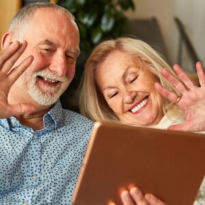An image of a man and woman seated together, both smiling and waving to someone they're chatting with on their tablet computer. Social Connections: The Power of Enhancing Your Well-Being as You Age - The Art of Aging Well
