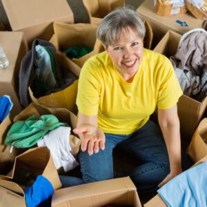 A middle-aged or senior woman, wearing a bright yellow tee shirt and jeans, seated in the middle of boxes and clothing donations as she smiles at the camera, her hand outstretched.