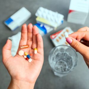 An image from the point of view of a person taking their medications; pills in one hand and a white pill held in the other hand. Below are a glass of water and an assortment of pills. Take your meds