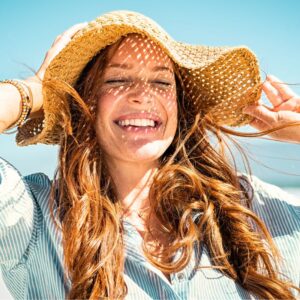 An image of a woman smiling with her eyes shut, wind blowing her hair, as she holds her straw hat down with her hands. Stay out of the Sun and The Art of Aging Well