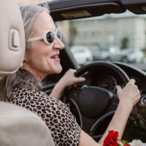 An image of a a woman wearing groovy white sunglasses and a leopard print top, smiling as she drives. Pick a car.