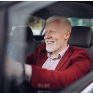 An image of a middle-aged man wearing a button up shirt and red sports jacket is smiling as he drives a car.