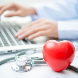 An image of a stethoscope and a red heart toy in the foreground of a doctor's hands typing on keyboard.