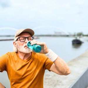 An image of a middle-aged man sitting beside a body of water, wearing a beige cap, black glasses, and an orange tee shirt taking a sip from his blue water bottle to stay hydrated.