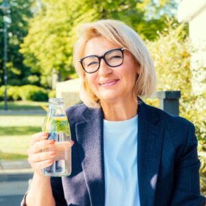 An image of a middle-aged woman smiling as she holds a bottle of water outdoors. Drink water to avoid UTIs
