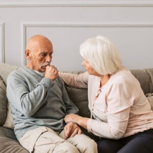 A seated man has a persistent cough and wheezing while his female companion comforts him.