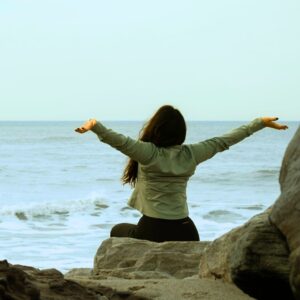 A person sitting on rocks near the ocean, arms in the air, feeling joy