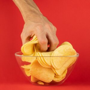 Hand grabbing potato chips from clear bowl with red background. Food addiction